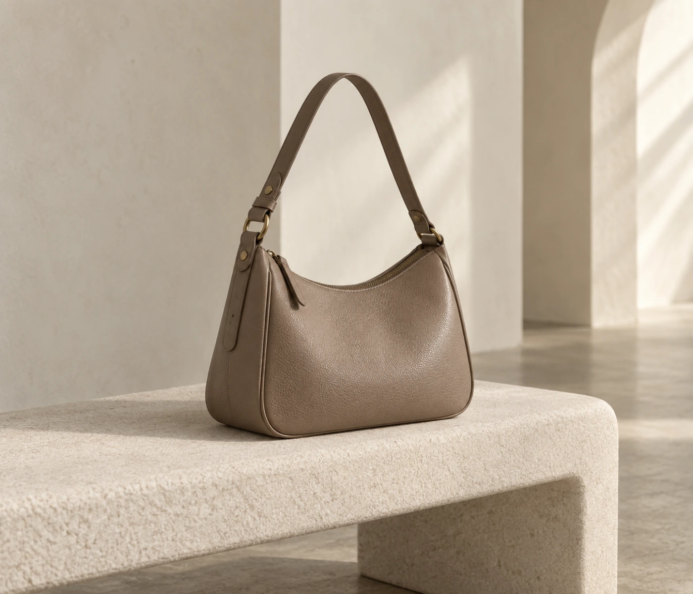 Taupe leather shoulder bag with a curved silhouette resting on an ivory upholstered bench.